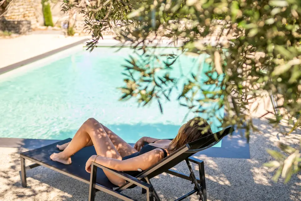 Une vacancière allongée sur un transat au bord de la piscine, à l'ombre d'un olivier